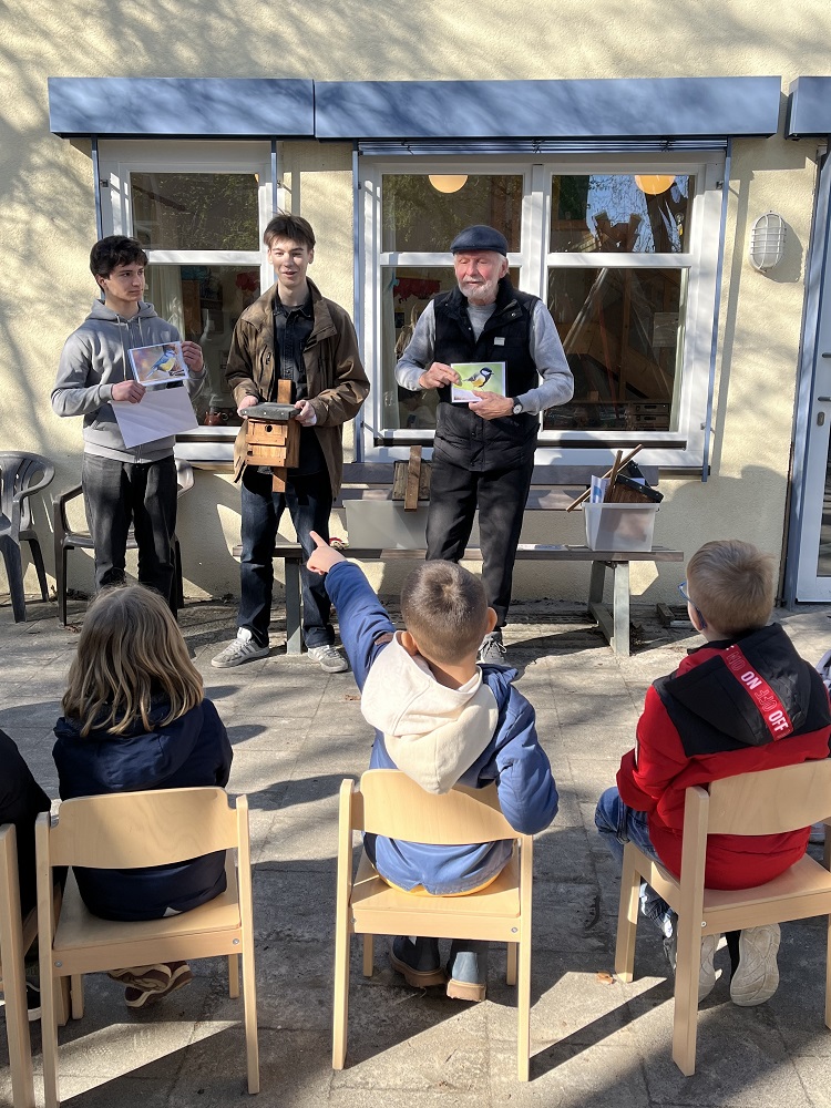 Foto: Kinder auf Stühlen vor Männern, die Nistkästen zeigen