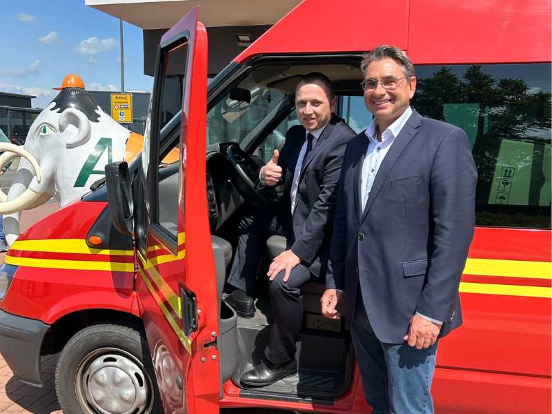 zwei Männer stehen an Fahrzeugtür eines roten Feuerwehrbusses, lächeln in die Kamera