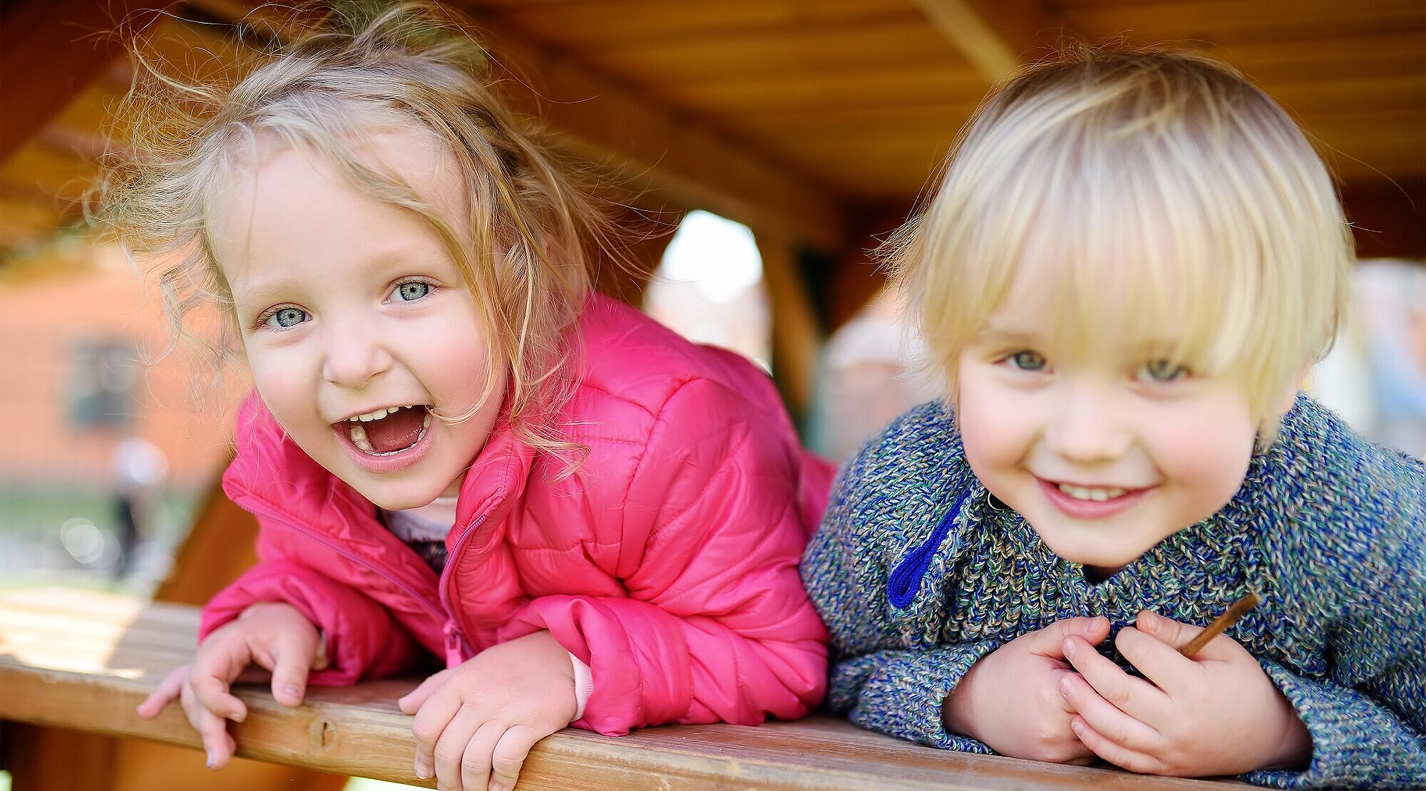 Foto: Zwei kleine Kinder schauen lachend aus einem Holzgerüst auf einem Spielplatz