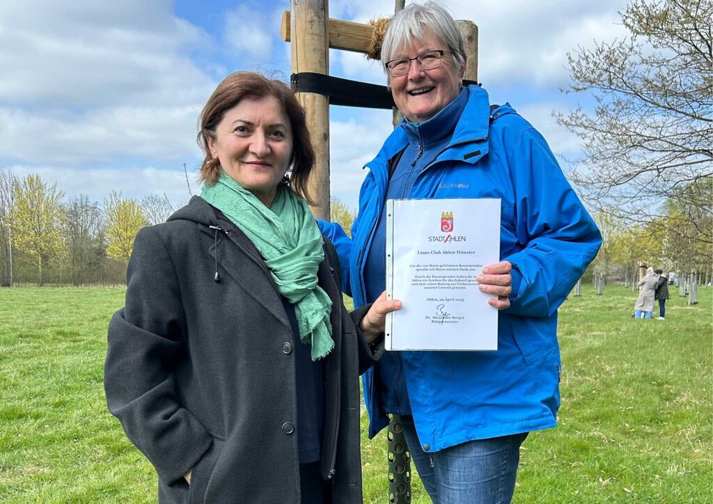 zwei Frauen stehen im Park vor einem frisch gepflanzten Baum mit Urkunde in der Hand