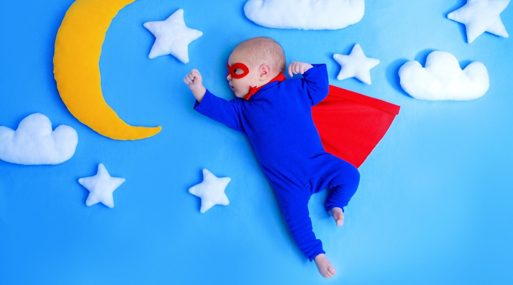 Foto: Baby im Superman-Kostüm liegt auf einer blauen Decke mit Wölkchen